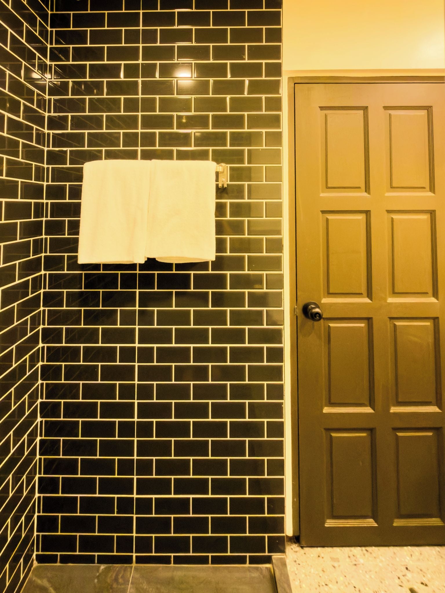 A bathroom with a black tiled wall, a white towel hanging on a rack, and a wooden door.