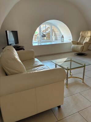 A living room with a sofa, armchair, and a glass coffee table. A television is on a small table to the left.