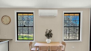 Et spiseområde med et bord og stole, to vinduer med udsigt til træer, og en airconditionenhed på væggen.