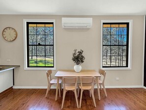 Ein Essbereich mit Tisch und Stühlen, zwei Fenster mit Blick auf Bäume und eine Klimaanlage an der Wand.