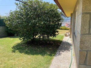 Um gramado bem cuidado com uma grande árvore ao centro, um muro de pedra à direita e uma vista desobstruída da região.