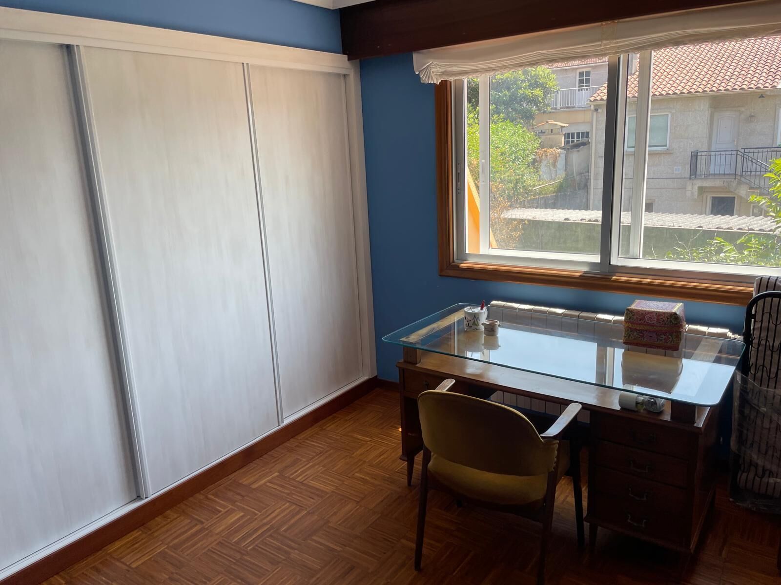 Una habitación con piso de madera, una mesa de vidrio, una silla, un ventanal con vista a edificios y un armario blanco.