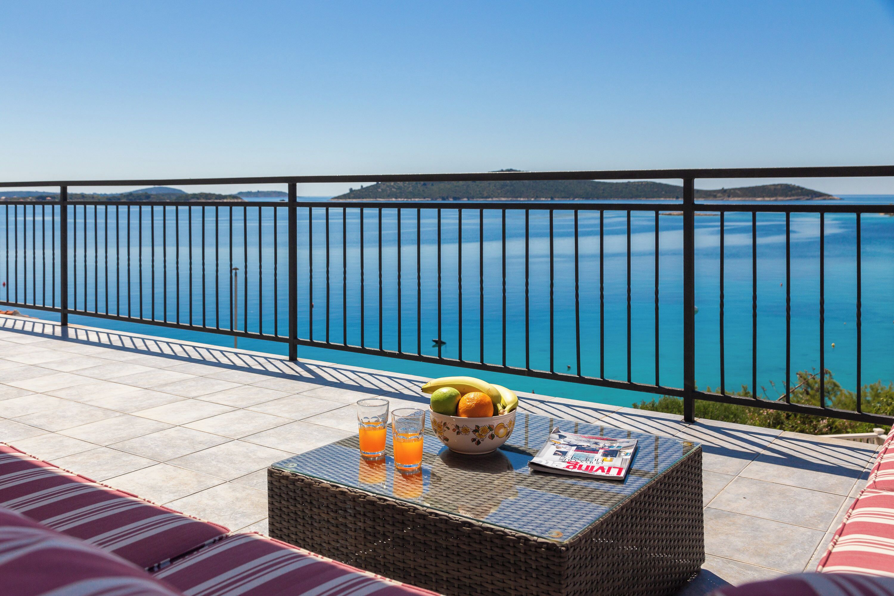 A balcony with a view of the sea, a wicker table with a bowl of fruit and glasses of orange juice, and striped cushions.