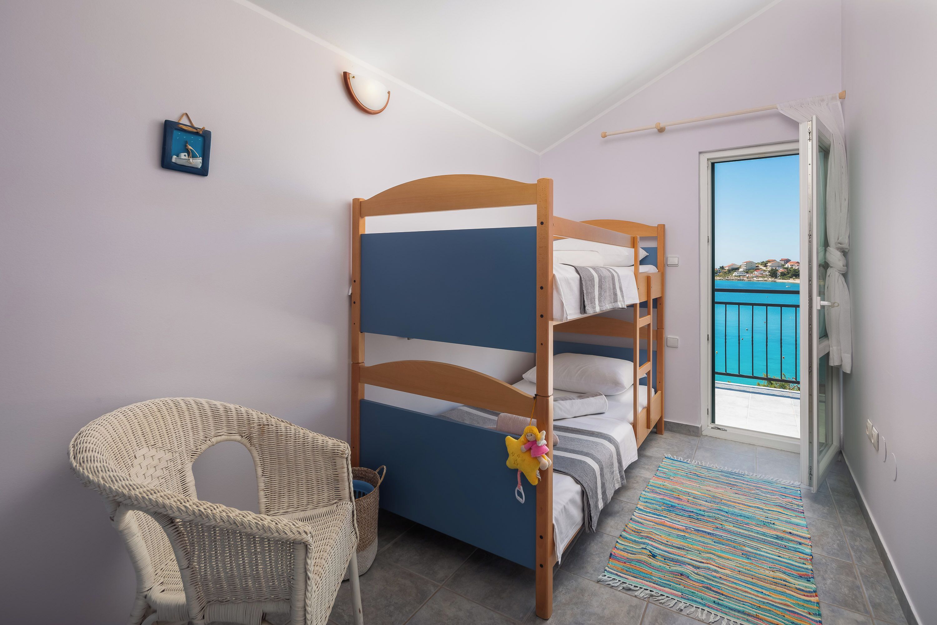 A room with a bunk bed, a wicker chair, a small table, and a view of the sea through a glass door.