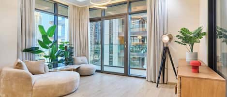 Una sala de estar moderna con un sofá curvo, una pequeña mesa de centro y una lámpara de pie. Hay ventanas grandes con cortinas, una planta en maceta y vistas de un paisaje urbano.