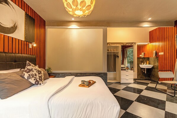 A modern bedroom with a large bed, a bedside table, a nightstand, a wall-mounted light fixture, and a view into the bathroom.