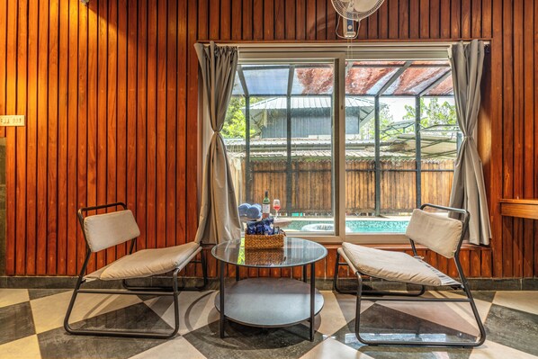 Una habitación con pared de paneles de madera, una mesa de vidrio con dos sillas, una ventana con cortinas y vista a una piscina exterior.