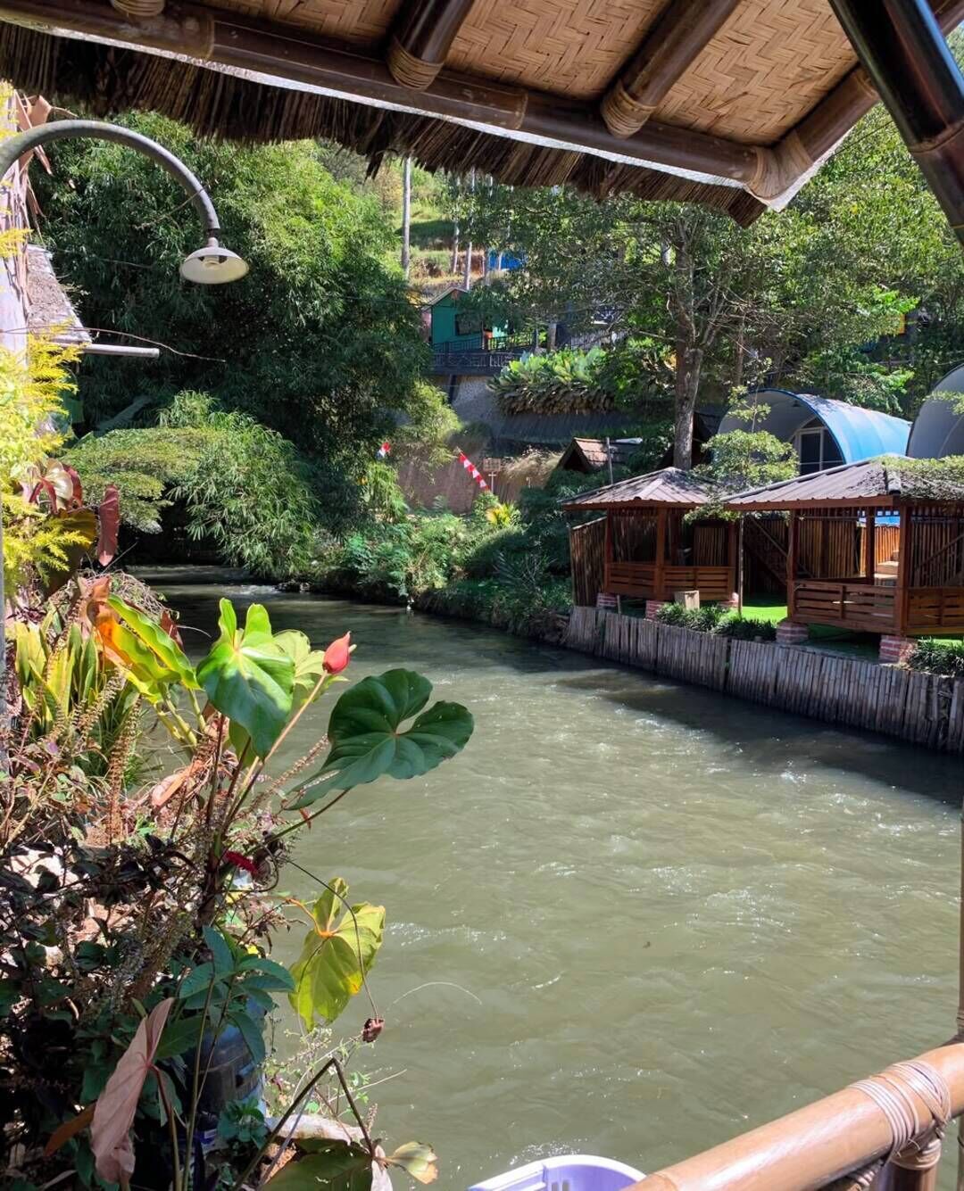 Une rivière traversée par un pont en bois, entourée d’une végétation luxuriante et bordée d’une construction à toit de chaume.