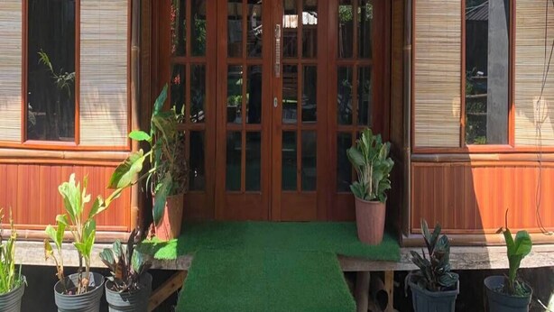 A wooden structure with a thatched roof, a central door with glass panels, and potted plants on the ground.
