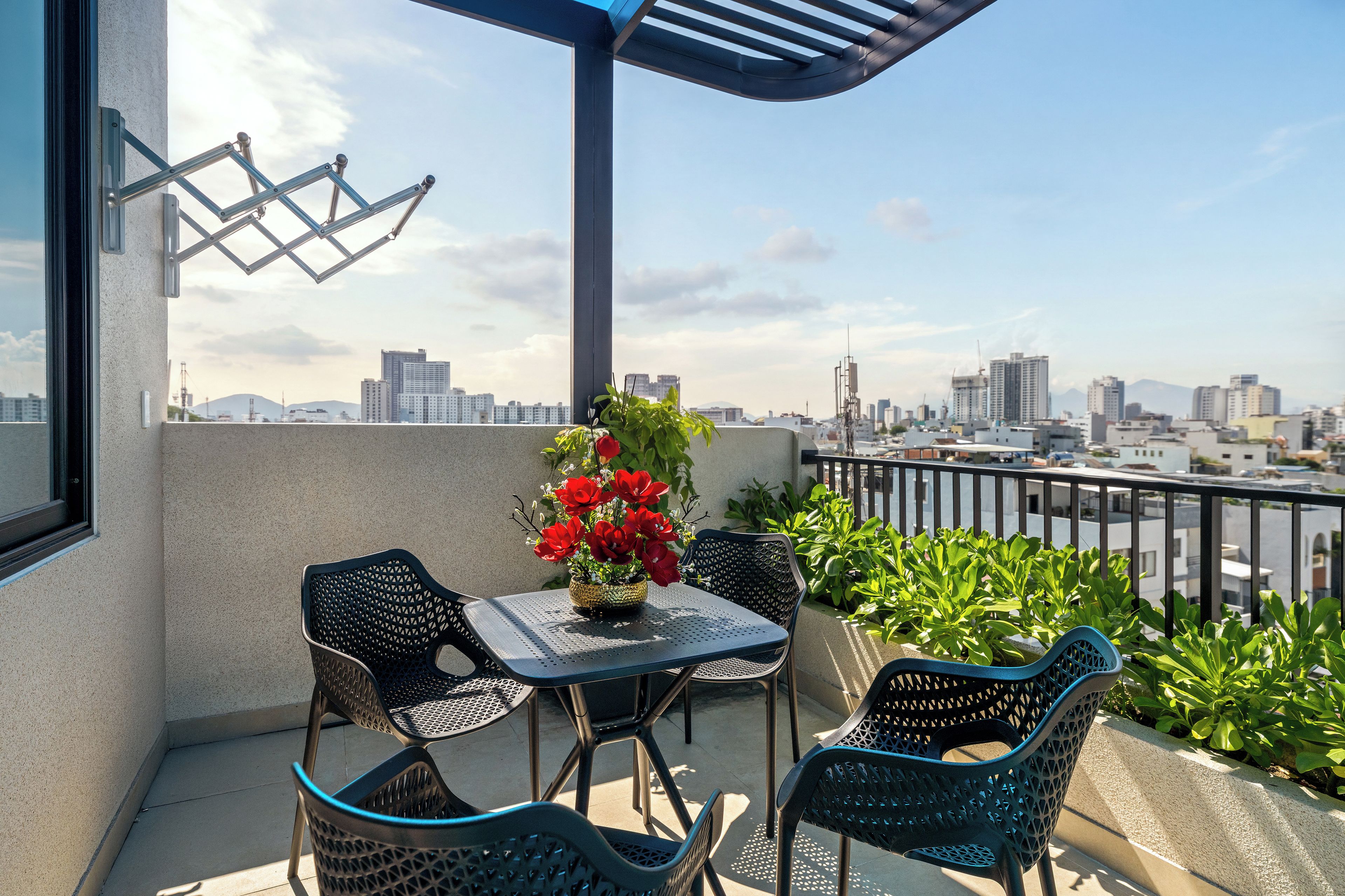 Uma varanda com mesa e cadeiras, um vaso com flores vermelhas e vista para a cidade.