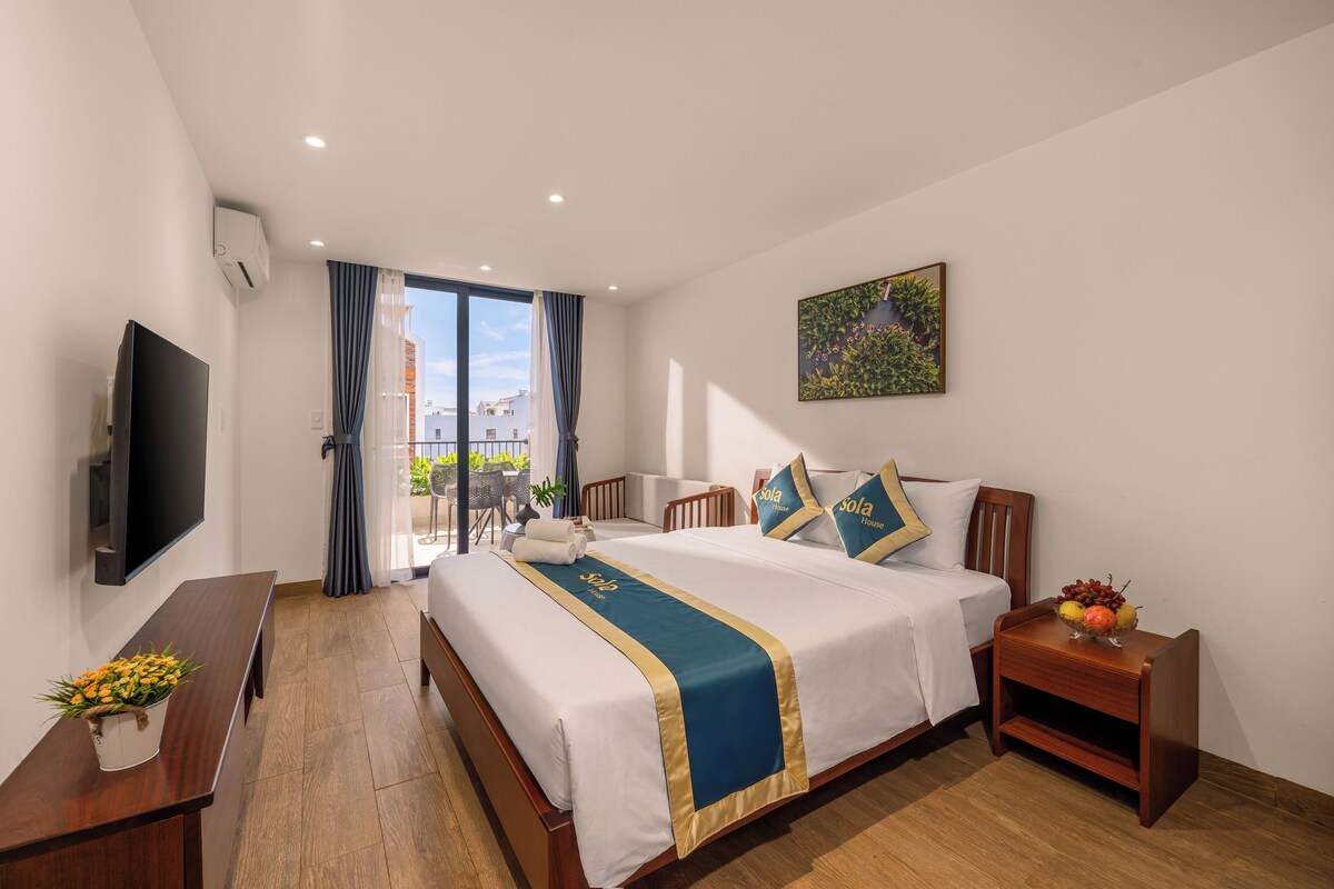 Quarto de hotel com cama, televisão, escrivaninha, cadeira, mesinha com frutas e varanda com mobiliário de exterior.