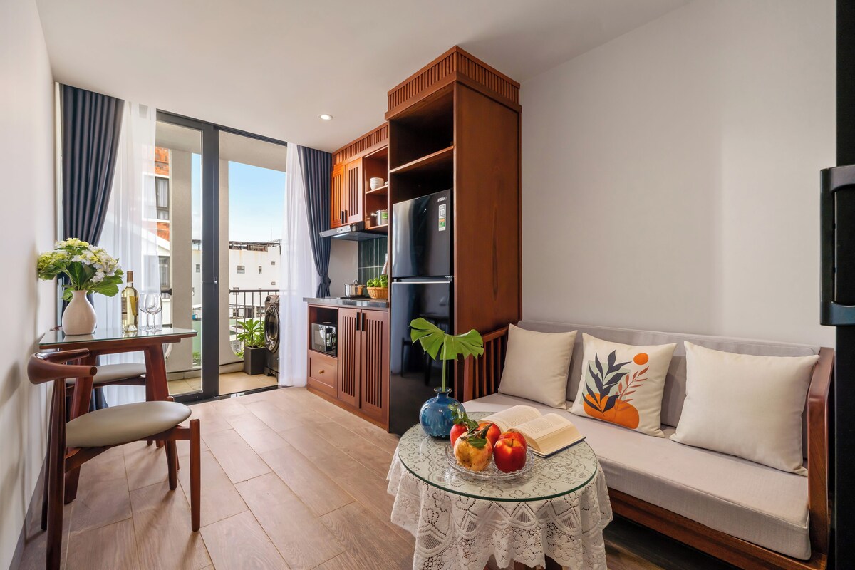 Uma sala de estar moderna com um sofá, uma mesa de centro com frutas e um livro, uma área de jantar com mesa e cadeiras, e um espaço de cozinha com armários e eletrodomésticos.