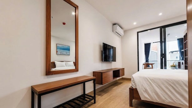 Una habitación de hotel moderna con piso de madera, un espejo grande, un televisor de pantalla plana, una mesita de noche y una cama con ropa blanca.