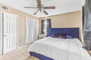 Un dormitorio con cama, ventilador de techo, mesita de noche y ventana con cortinas.
