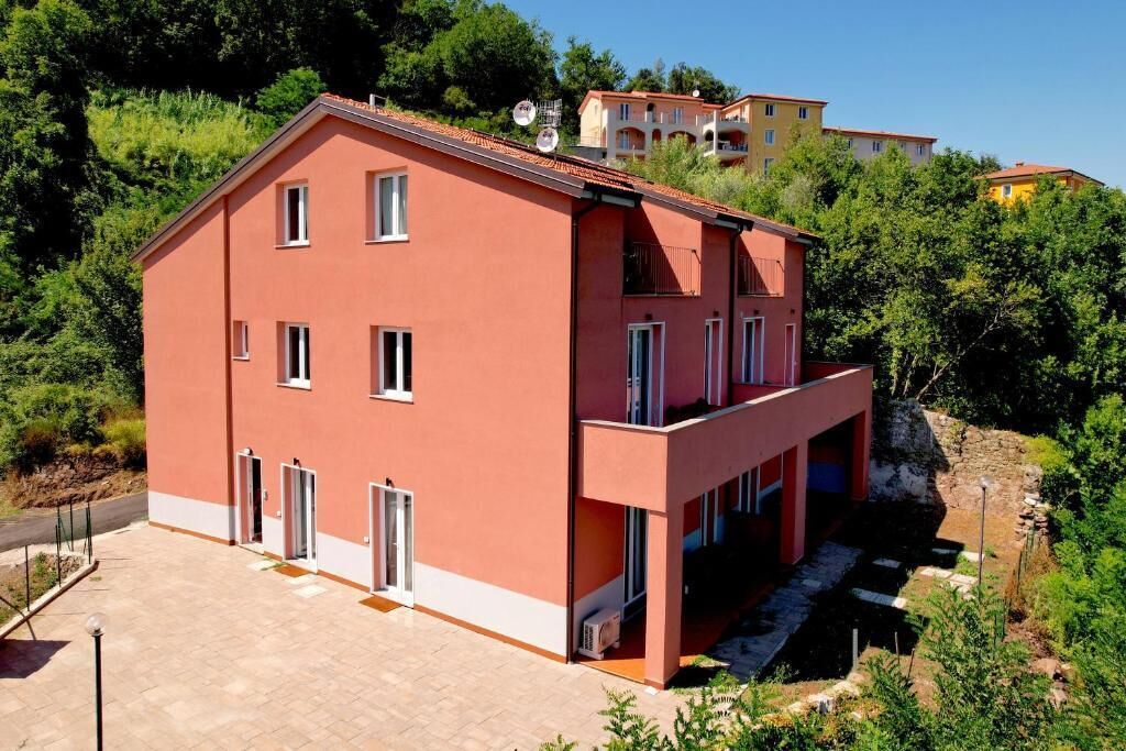 A two-story building with a flat roof, a balcony on the upper floor, and a paved entrance area.