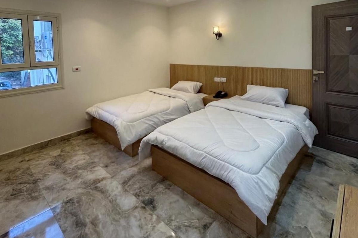 A hotel room with two beds, a wooden headboard, a nightstand, a wall-mounted light, and a door.