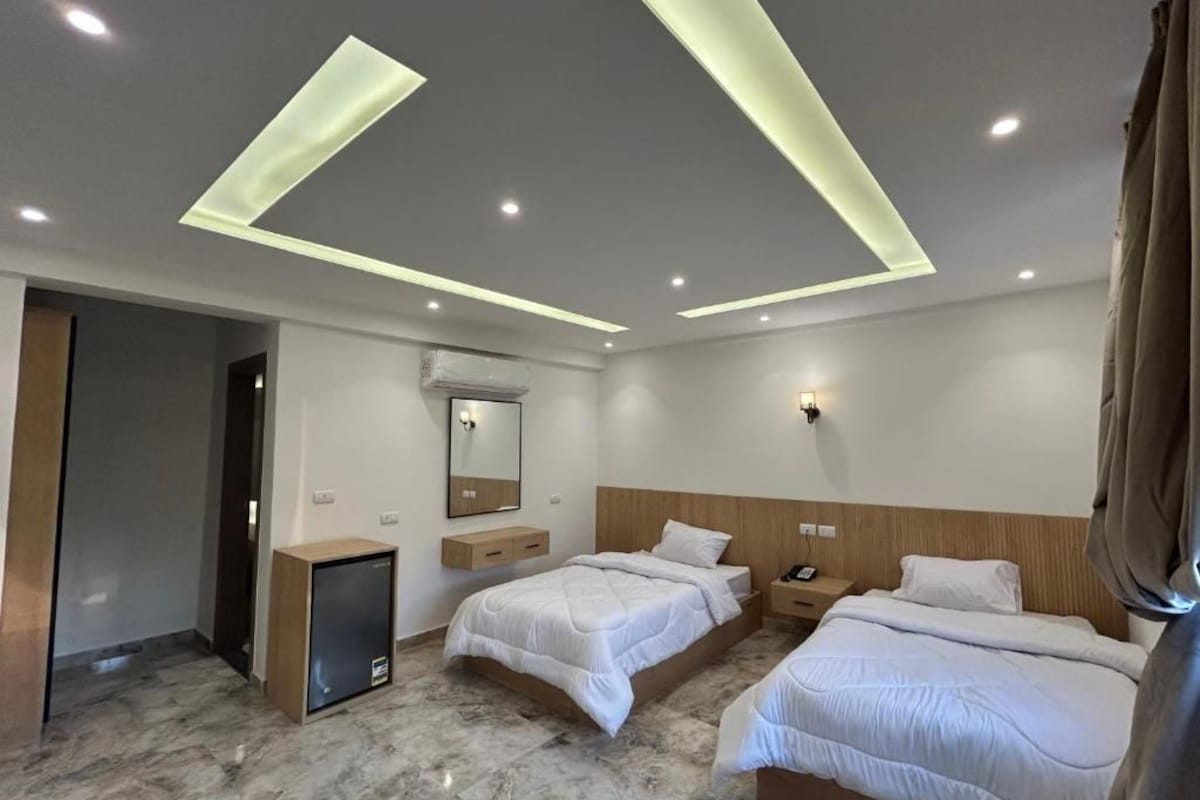 A hotel room with two beds, a desk, a mirror, and recessed ceiling lighting.