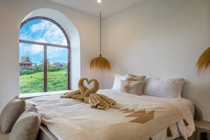 A hotel room with a large arched window, a bed with a heart-shaped towel arrangement, and a view of greenery and buildings outside. - 3BR Private Pool Villa with Rooftop in Pererenan Near Canggu (Pererenan)