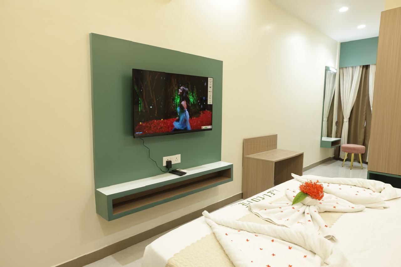 Een hotelkamer met een flatscreen televisie aan de muur, een bed met een keurig opgevouwen handdoek en een bloem, en een klein nachtkastje.