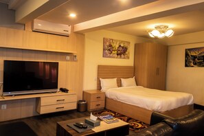 Une chambre d’hôtel dotée d’une grande télévision à écran plat, d’un lit avec du linge de lit blanc, d’une armoire en bois, d’une petite table de chevet et d’un luminaire au plafond.