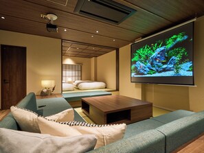 A modern hotel room with a large screen displaying an aquarium, a sofa, a coffee table, and a bed with a view of the room.