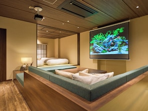 A modern hotel room with a large bed, a seating area, a television displaying an aquarium, and a wooden ceiling.