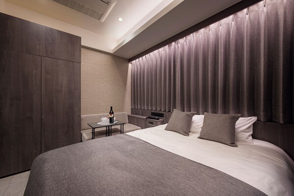 A modern hotel room with a large bed, a bedside table, a small table with a bottle and glasses, and a wardrobe.