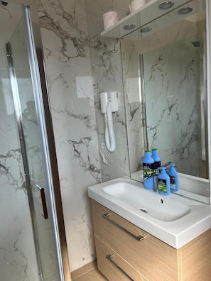 A bathroom with a glass shower enclosure, a marble wall, a white sink with a mirror above it, and toiletries on the sink. - Two-Bedroom Family Apartment (Venezia)