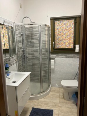 A bathroom with a glass-enclosed shower, a white sink, a toilet, and a window with a colorful pattern. - Two-Bedroom Family Apartment (Venezia)