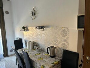 A dining area with a table set for two, a wall clock, a television mounted on the wall, and a patterned wallpaper. - Two-Bedroom Family Apartment (Venezia)