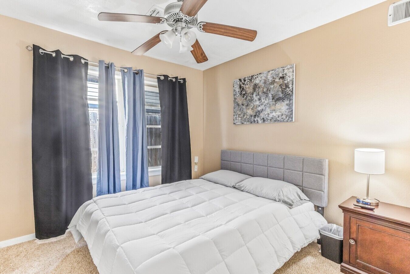 A bedroom with a bed, curtains, a ceiling fan, a nightstand, and a wall art piece.