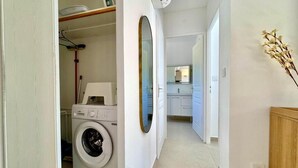 A laundry room with a washing machine, a mirror, and a shelf with a drying rack and ironing board. - ALBIZIA Residence: 1-Bedroom Apartment with Spa, Pool, and Parking - Renovated in 2026 (Gréoux-les-Bains)