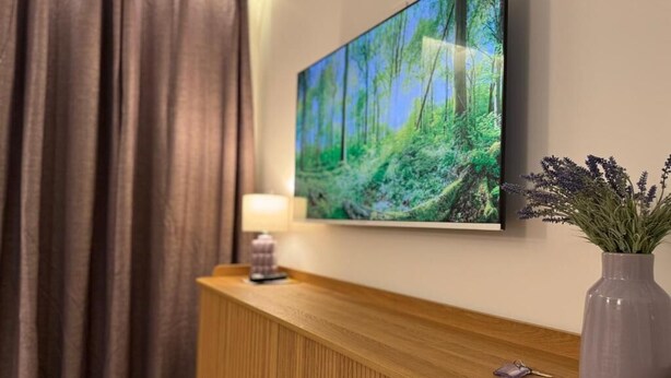 A hotel reception area with a wooden counter, a flat-screen TV displaying a forest scene, a vase with purple flowers, and a lamp. - ALBIZIA Residence: 1-Bedroom Apartment with Spa, Pool, and Parking - Renovated in 2026 (Gréoux-les-Bains)