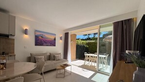 A modern living room with a sofa, coffee table, and dining table. There is a painting on the wall and a view of the garden through the sliding glass door. - ALBIZIA Residence: 1-Bedroom Apartment with Spa, Pool, and Parking - Renovated in 2026 (Gréoux-les-Bains)