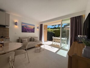 A modern living room with a sofa, coffee table, and dining table. There is a painting on the wall and a view of the garden through the sliding glass door. - ALBIZIA Residence: 1-Bedroom Apartment with Spa, Pool, and Parking - Renovated in 2026 (Gréoux-les-Bains)