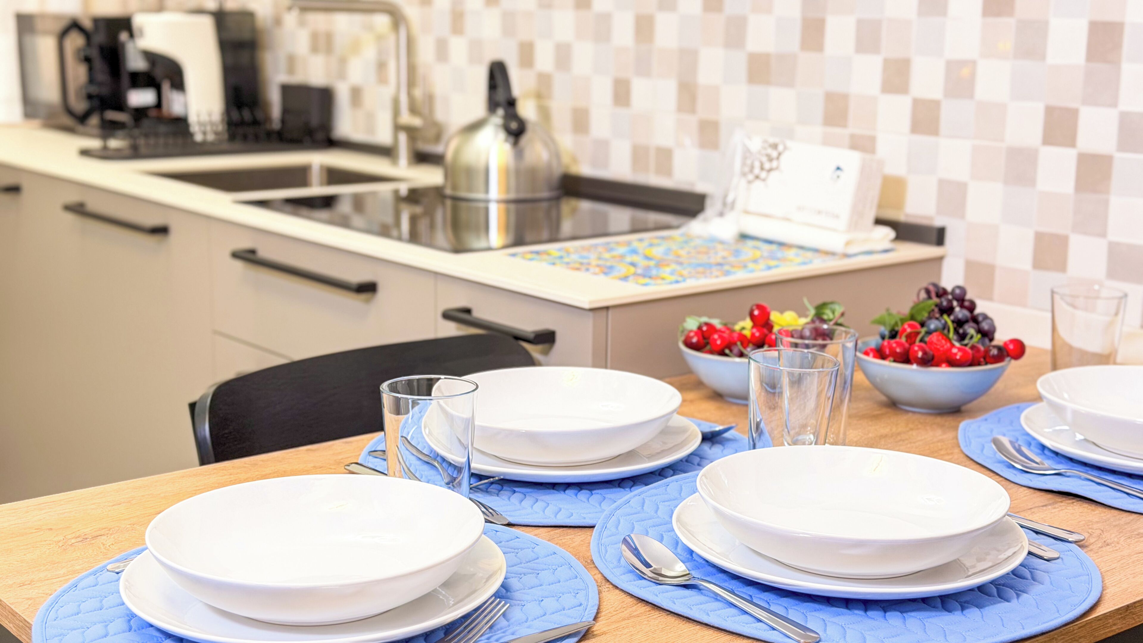 A dining table set with plates, glasses, and cutlery, bowls of fruit, and a kettle in a kitchen.