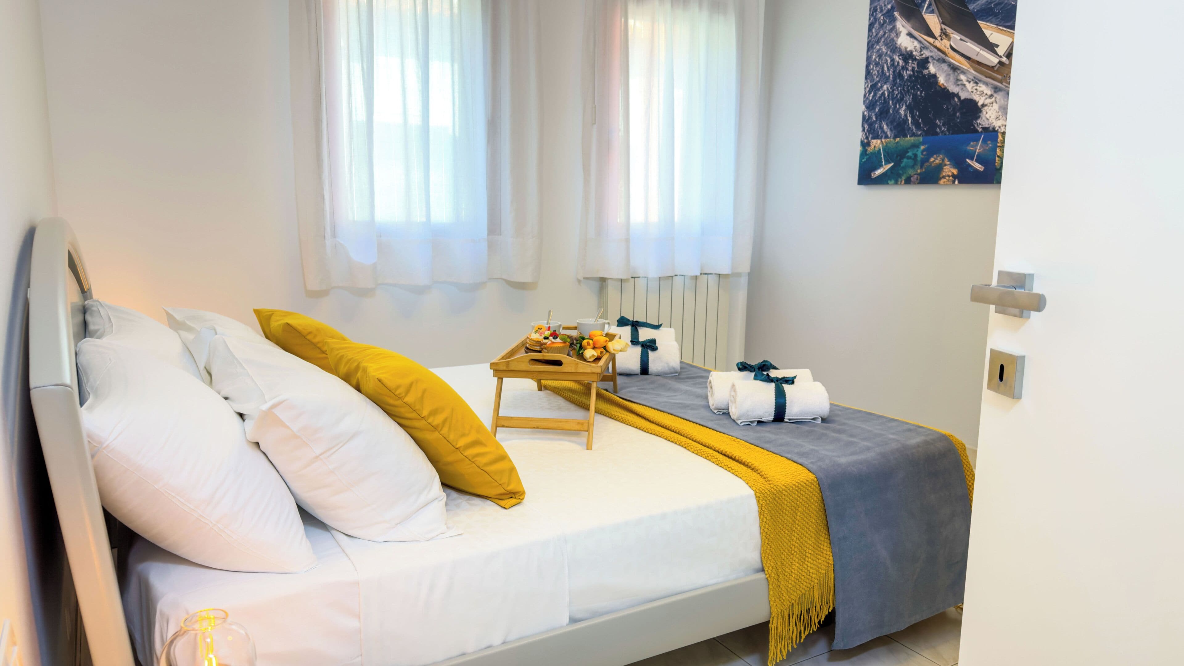 Una cama bien hecha con ropa de cama blanca y amarilla, una bandeja con cosas para el desayuno y un cuadro de un barco en la pared.