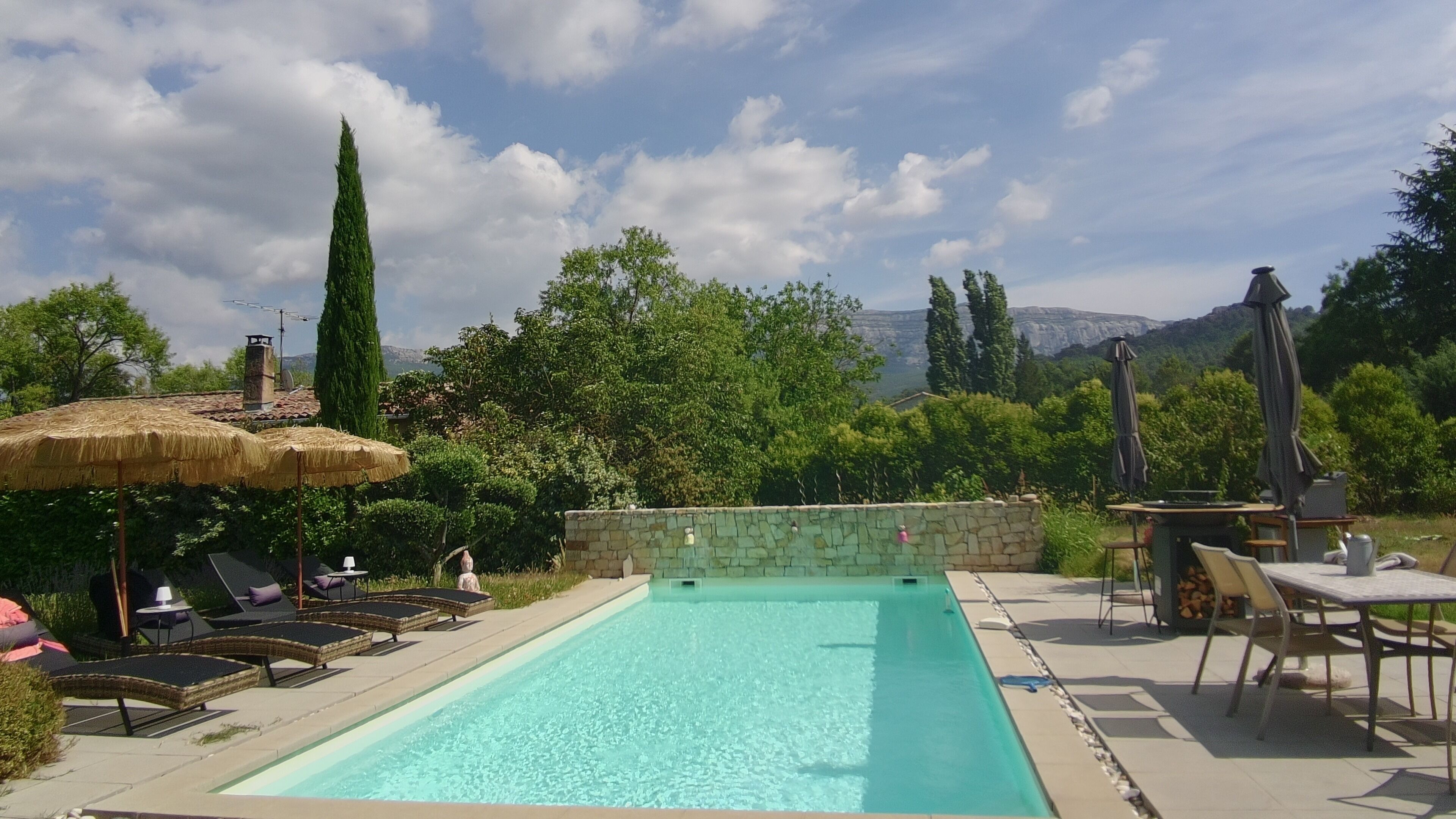 Piscina con sillones de descanso y sombrillas, rodeada de vegetación y un muro de piedra.
