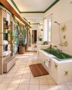 A bathroom with a large green bathtub, a shower, and a wall with a tropical mural. There are shelves with towels and plants, and a wooden bench. - Maison Sarava – Charming apartment with balconies and spacious rooms (Espira-de-l'Agly)