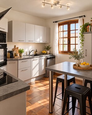 Una cuina moderna amb armaris blancs, un esbandidor d'acer inoxidable i una zona de dinar amb taula i tamborets.
