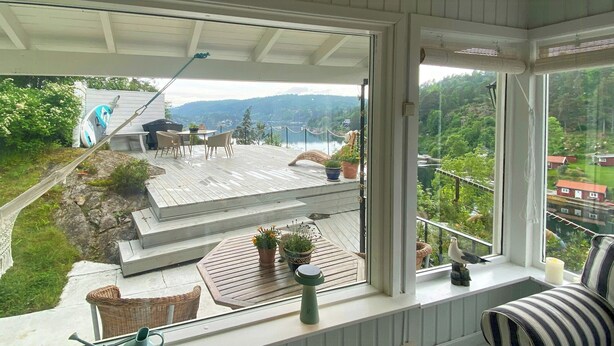 A room with a large window overlooking a deck with outdoor furniture and a view of a lake and mountains.