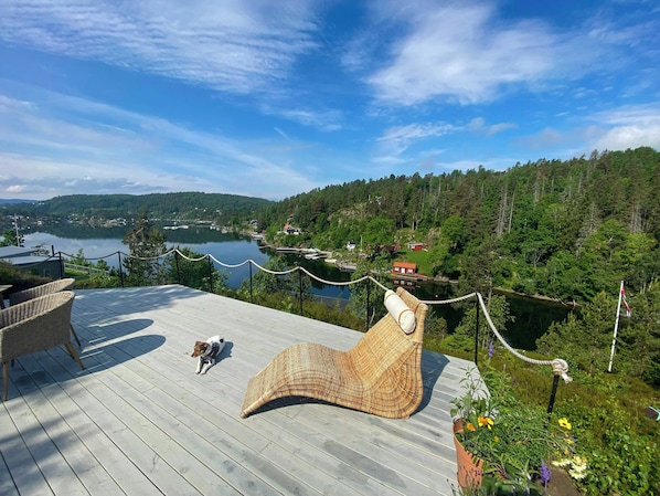 Terraza de madera con mobiliario de mimbre que da a un lago y colinas boscosas.