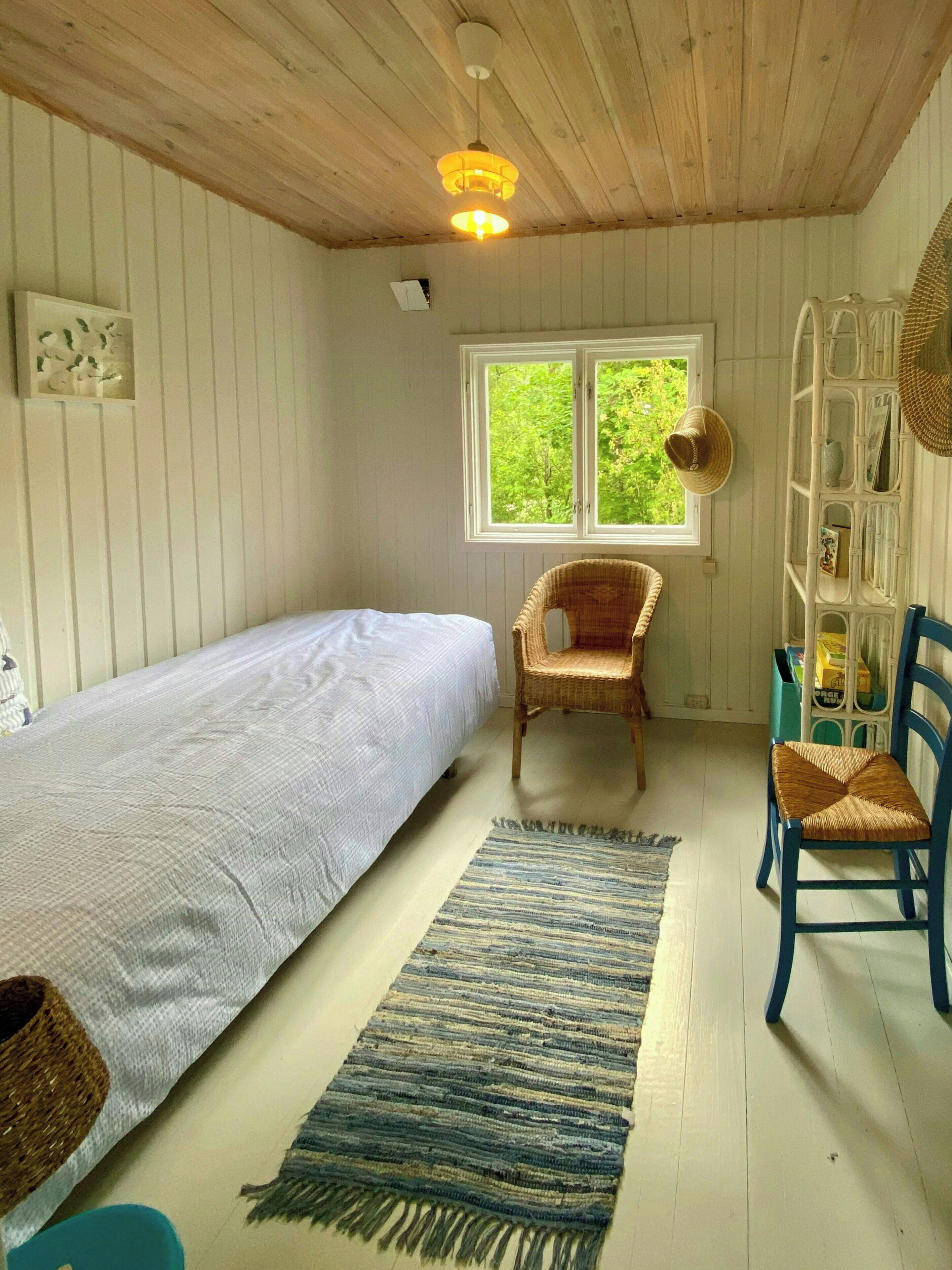 Une chambre comprenant un lit simple, une chaise, une petite table, une fenêtre donnant sur un espace verdoyant et une étagère contenant divers objets.