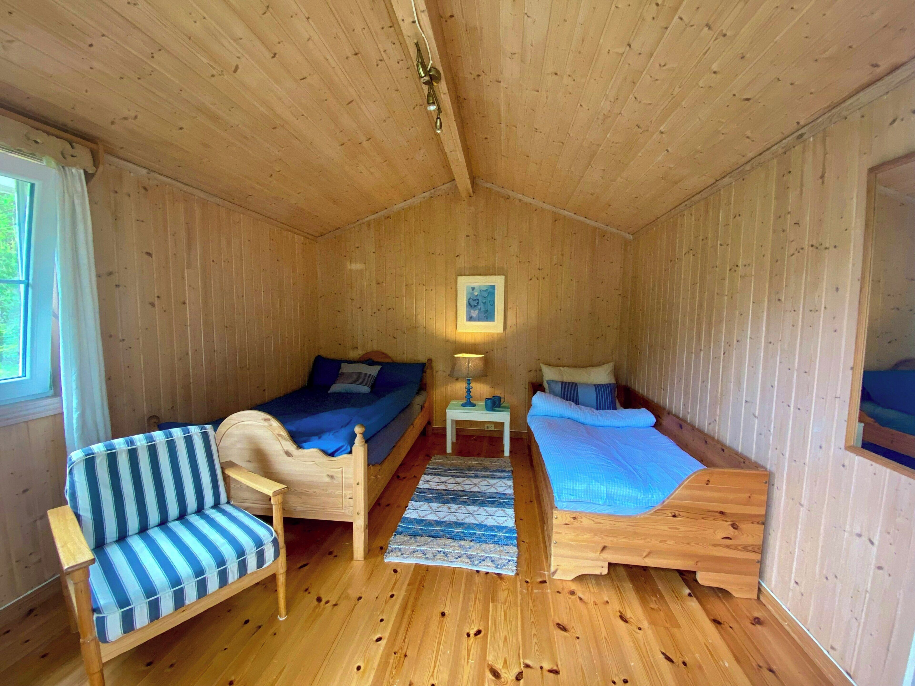 A wooden cabin with two single beds, a striped sofa, a small table with a lamp, and a window with curtains.