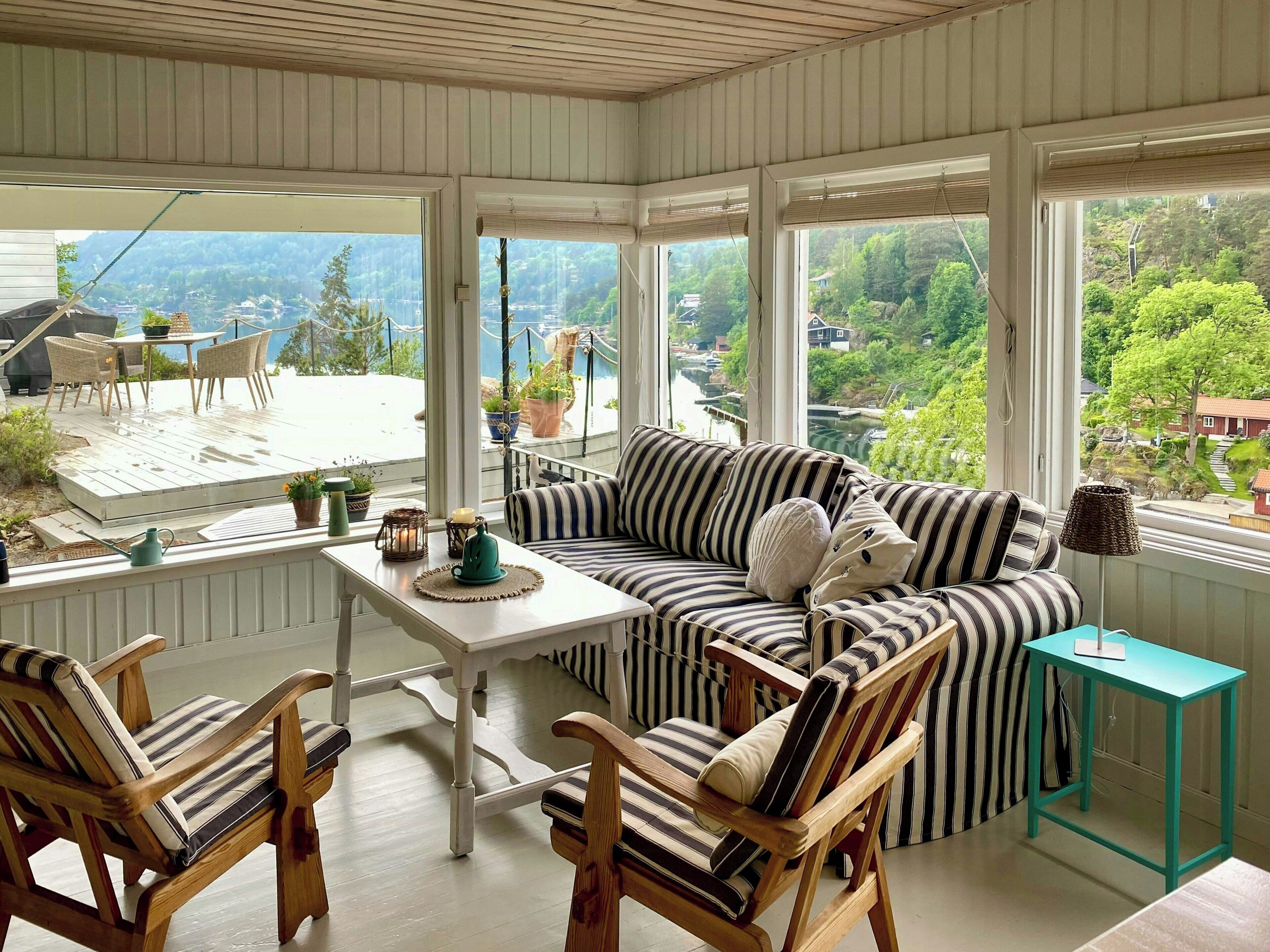 A sunlit room with a view of a river and greenery, featuring a striped sofa, wooden chairs, and a small table with decorative items.