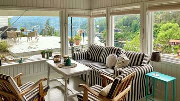 A sunlit room with a view of a river and greenery, featuring a striped sofa, wooden chairs, and a small table with decorative items.