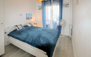 Een slaapkamer met een bed, nachtkastje met lamp, een ventilator en een raam met gordijnen.
