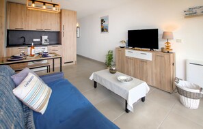 A modern living room with a blue sofa, a small table, a television, and a kitchen area with wooden cabinets and a coffee machine. - 1 bedroom lovely apartment in Théoule sur Mer (Théoule sur Mer)