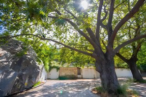 A tree-lined courtyard with a paved area, a stone wall, and a building in the background. - Hillside Hideaway mountain villa with views, shared terrace, and private garden (Skiadas)