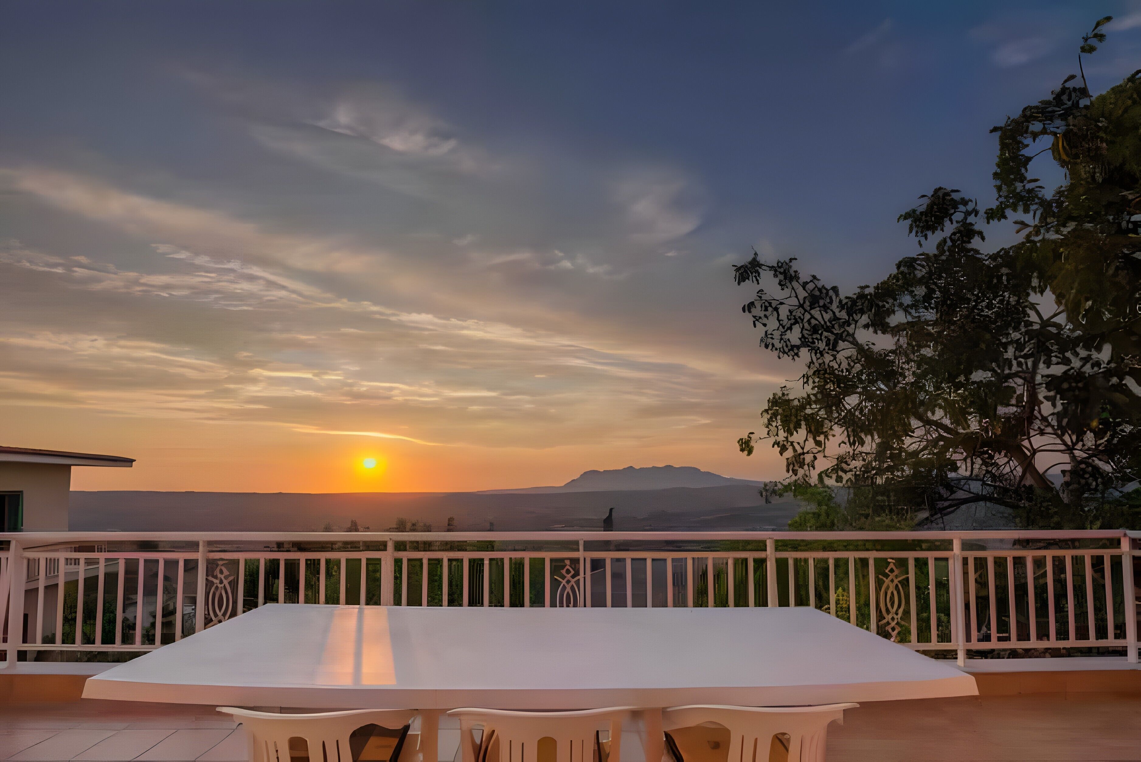 A balcony with a white table and chairs, overlooking a sunset with mountains in the distance.